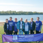 4 women and 3 men wearing I walk to end pancreatic cancer t-shirts, standing in front of a Purple Stride Iowa 2019 banner