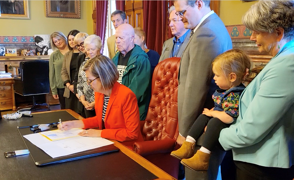 About a dozen people stand behind Governor Kim Reynolds' desk as the Governor signs the Gail Orcutt School Radon Safety Bill.