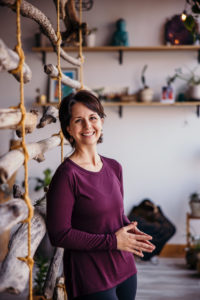 Melissa, a woman with short brown hair, wears a long-sleeve purple shirt, and smiles warmly at the camera in front of a natural wood art display and potted plants in her yoga studio.