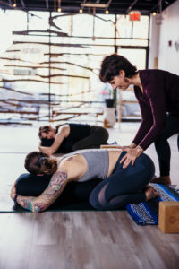 Melissa helps a yoga client position their hips correctly. Several other yoga students practice in a natural-looking and well-lit yoga studio.