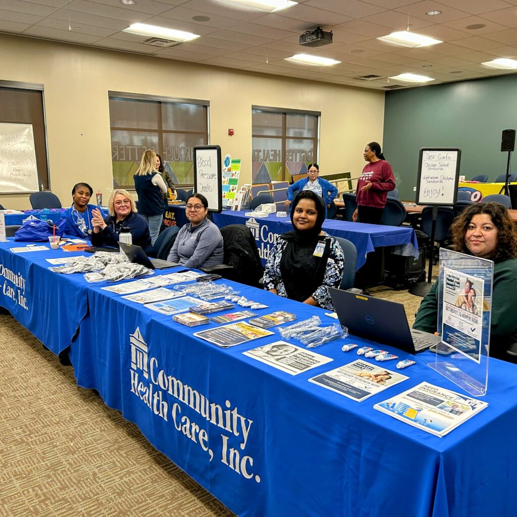 Several employees of Community Health Care, Inc., sit behind a table at a community event with cancer screening information on their table.