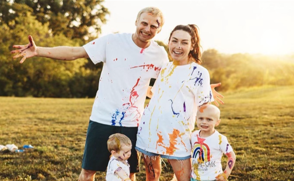 A young couple and their two young children (Natalie and her family) pose in a field at sunset, wearing white t-shirts covered in handprints of colorful paint.